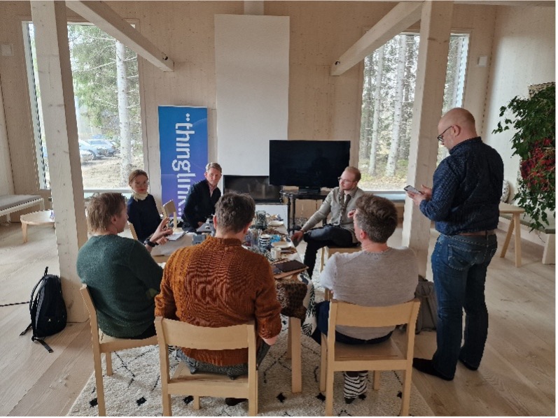 Seitsemän henkilöä istuu ja seisoo keskustelupiirissä valoisan, puupintaisen huoneen keskellä. Keskellä on sohvapöytä, jolla on kahvia ja tarjoiluja. Taustalla näkyy ThingLinkin pystybanneri, suuri televisio ja takka. Huoneessa on suuret ikkunat, joista näkyy metsäinen maisema. Oikealla seisoo mies katsomassa puhelintaan, muut osallistujat istuvat ja keskustelevat.